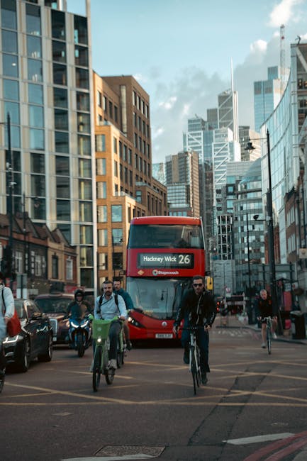 In the foreground, a group of people are riding bicycles through a busy city street lined with modern and historic buildings in Hackney, London. Some cyclists carry backpacks or bags, and they are rid