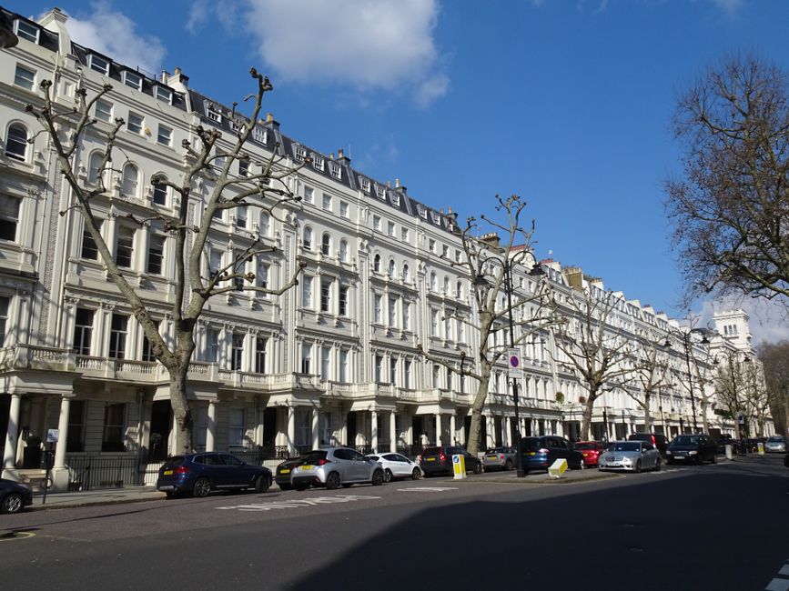 An exterior city street scene near London Fields featuring a multi-storey residential building with beige brickwork, white window frames, and a rounded corner turret, surrounded by leafless trees indi